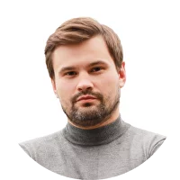 A man with short brown hair and a beard wearing a gray turtleneck sweater, looking directly at the camera against a white background.