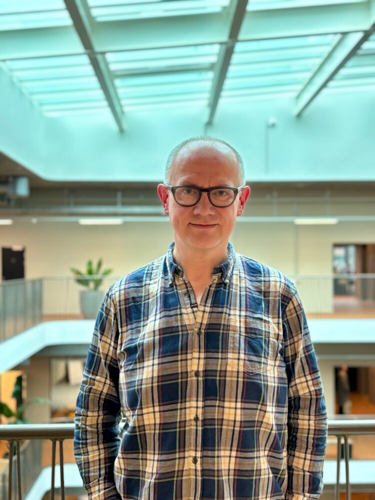 A man wearing glasses and a plaid shirt stands indoors in a modern building with large windows and natural light, reflecting the innovative environment at Visma.