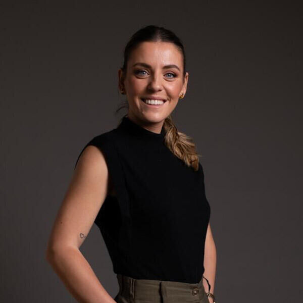 Marinela Profi A woman with light brown hair in a ponytail, wearing a sleeveless black top and khaki pants, stands smiling against a plain dark background.