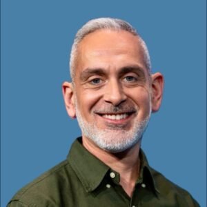 A middle-aged man with short gray hair and a beard, wearing a green button-up shirt, smiling in front of a solid blue background.