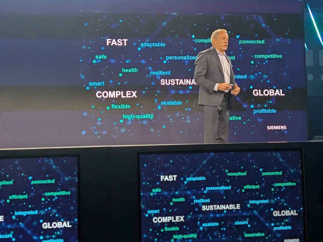 A man in a suit stands on stage in front of a screen displaying words like "FAST," "SUSTAINABLE," "COMPLEX," and "GLOBAL" with the Siemens logo.