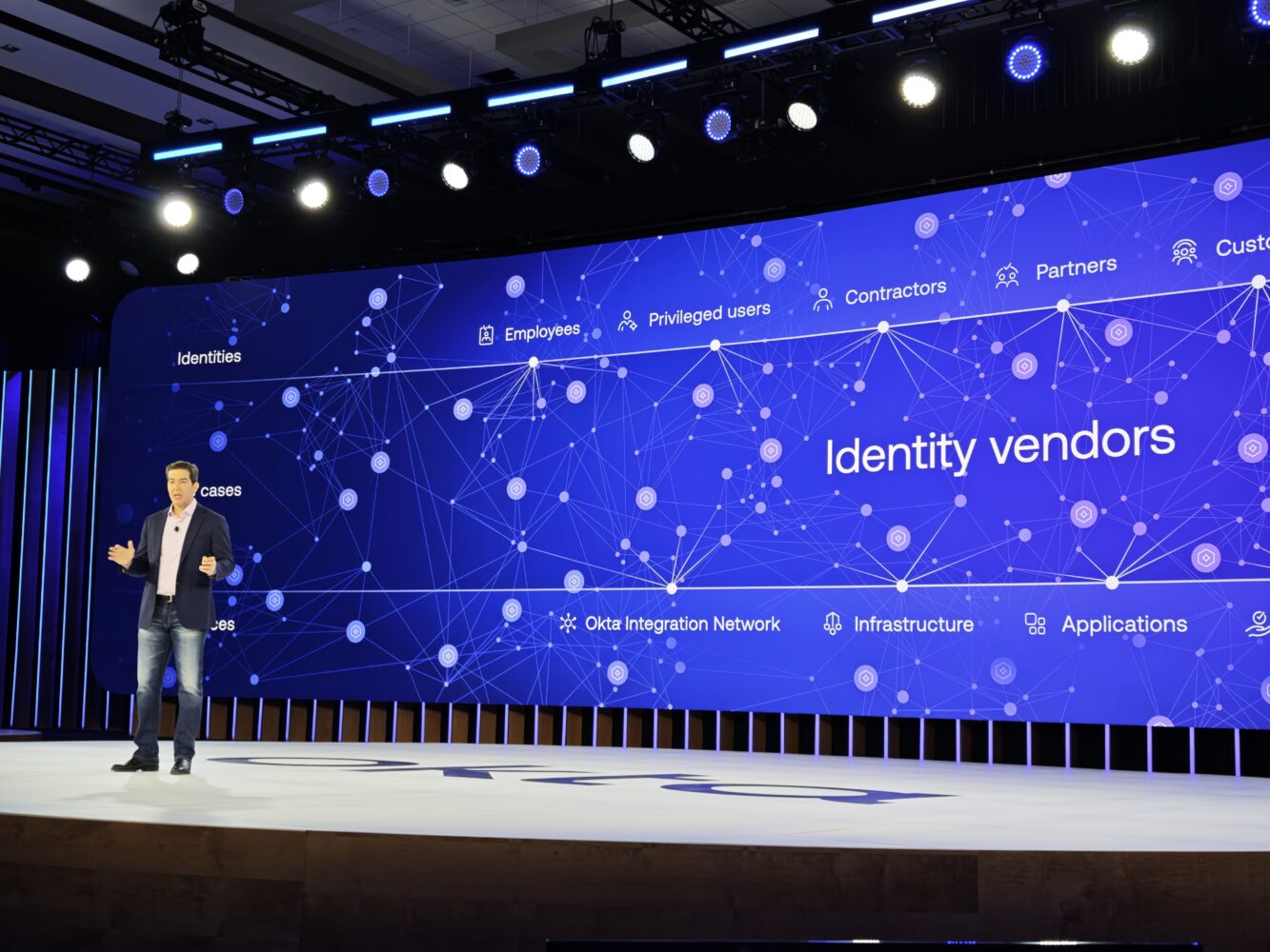 A man stands onstage in front of a large screen displaying a network diagram with the text "Identity vendors," related terms, Okta, and various icons.