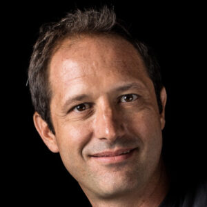 A man with short dark hair and a slight smile, wearing a black shirt, is pictured against a black background.