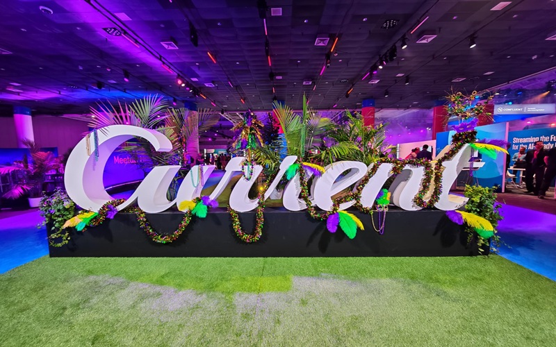 c8357078-42f4-4780-a860-88424e580c03 Large white "Current" sign decorated with colorful beads, feathers, and plants is displayed indoors on green artificial grass, under purple and blue lighting.
