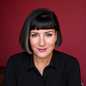 A woman with a short black bob hairstyle and bangs, wearing a black collared shirt, smiles calmly in front of a solid dark pink background.