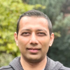 A man with short dark hair and a grey zip-up hoodie stands outdoors in front of blurred green foliage.