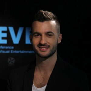 A man with short brown hair and light facial hair is smiling, wearing a black blazer over a white shirt, with a dark background and partially visible text.