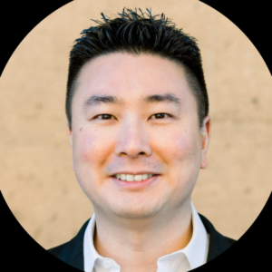 A man with short black hair, wearing a white shirt and dark blazer, smiles at the camera against a beige background.