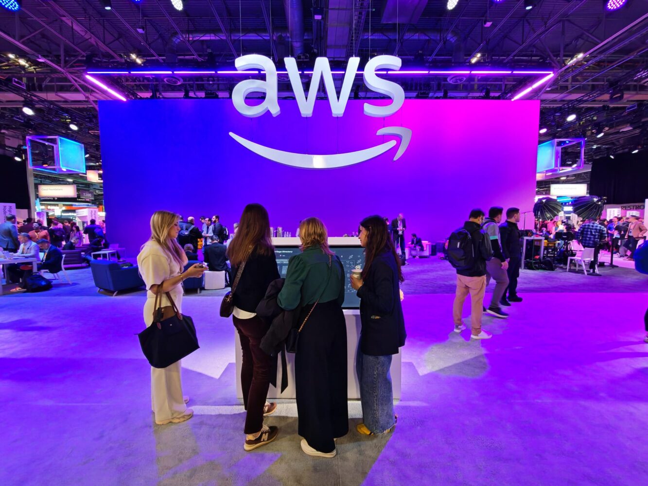 People gather around a display booth under a large illuminated "AWS" sign at a technology event with purple and blue lighting.