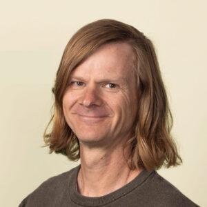 A man with shoulder-length light brown hair wearing a brown sweater stands against a plain light background, smiling at the camera.