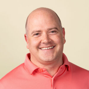 A smiling man with a shaved head wearing a coral-colored polo shirt, posed against a plain light background.