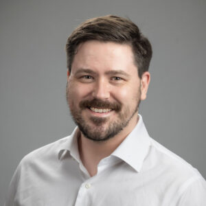 A man with short dark hair and a beard, wearing a white button-up shirt, smiling in front of a plain gray background.