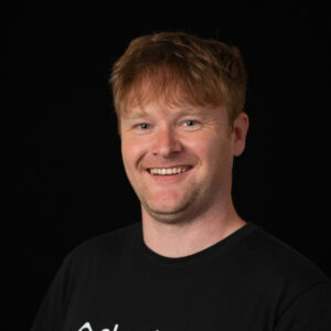 A person with light brown hair smiles at the camera, wearing a black t-shirt with the word "cloudsmith" printed on it, against a plain black background.