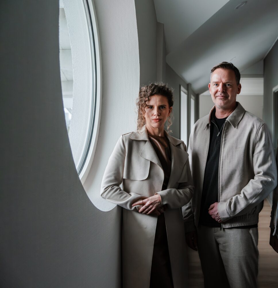 A woman and a man stand indoors near a round window, both wearing light-colored jackets and looking at the camera in what appears to be a stylish Yenlo-inspired setting.