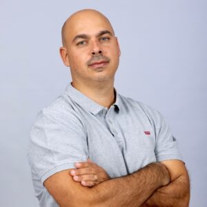 A man with a shaved head and mustache wearing a light gray polo shirt stands with arms crossed against a plain light gray background.