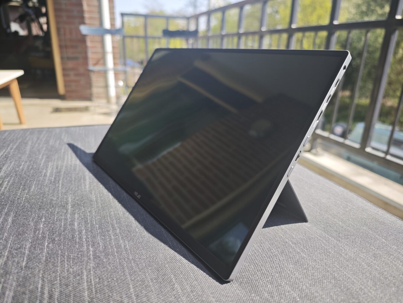 An ASUS ZenScreen OLED MQ16FC with a built-in kickstand is positioned on a table outdoors, with a balcony railing and greenery visible in the background.