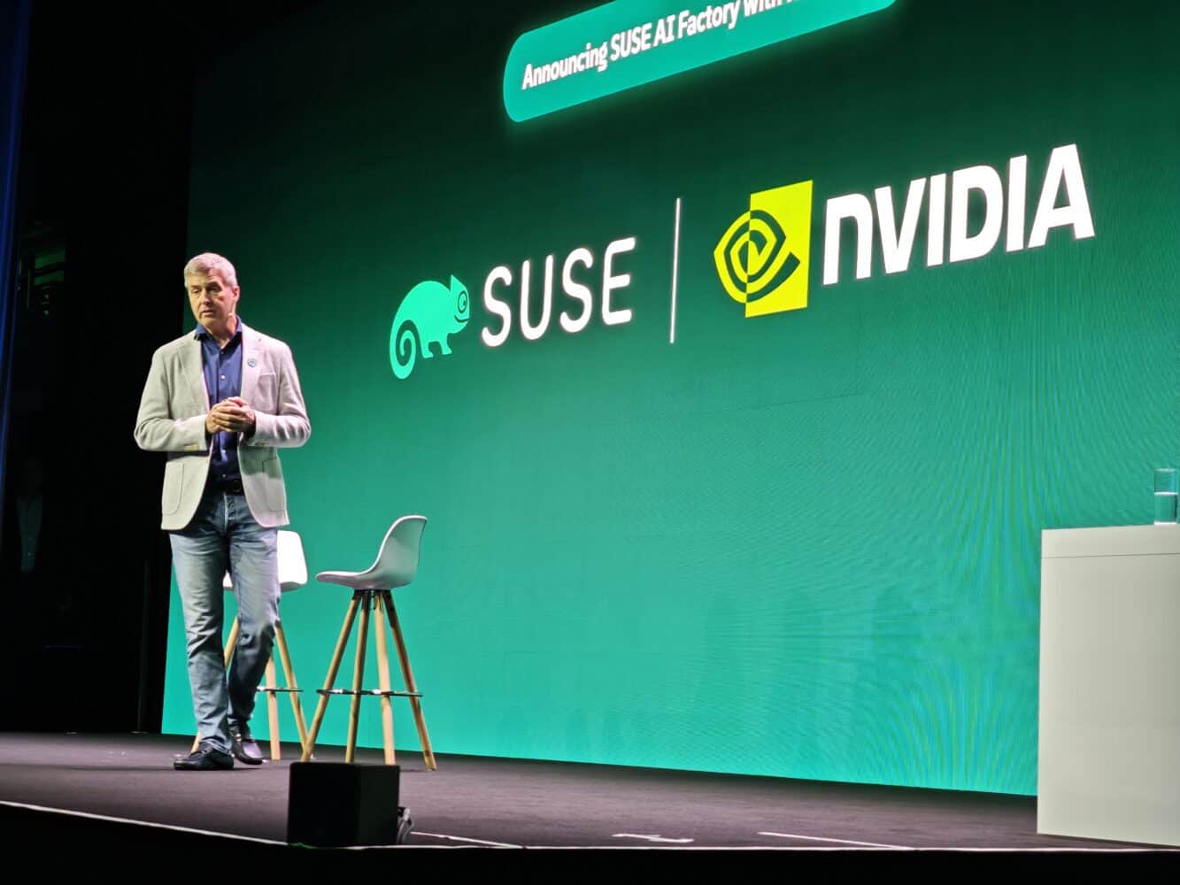 30316 A man stands on stage at SUSECON in front of a green screen displaying SUSE and NVIDIA logos, announcing the sovereign SUSE AI Factory with NVIDIA.