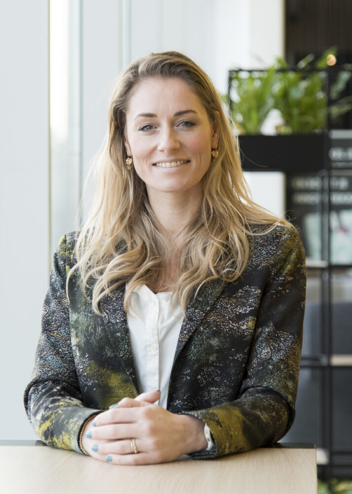 A woman with long blonde hair, wearing a patterned blazer and white blouse, sits at a table with her hands folded, smiling at the camera. There are plants and shelves in the background.