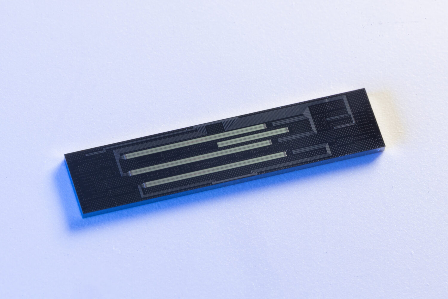 A rectangular black electronic chip, possibly a Universal Quantum Switch, with metallic lines sits on a white surface, illuminated with blue-tinted light.
