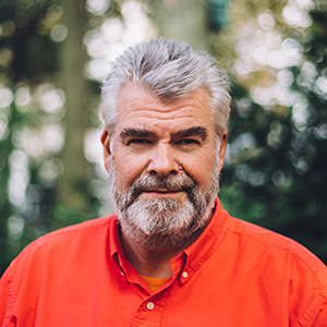 A middle-aged man with gray hair and a beard wearing an orange shirt stands outdoors with trees in the blurred background.