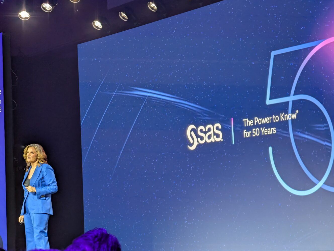 A woman in a blue suit stands on stage next to a large screen showcasing the SAS logo and the text "The Power to Know for 50 Years," celebrating SAS's legacy of innovation.