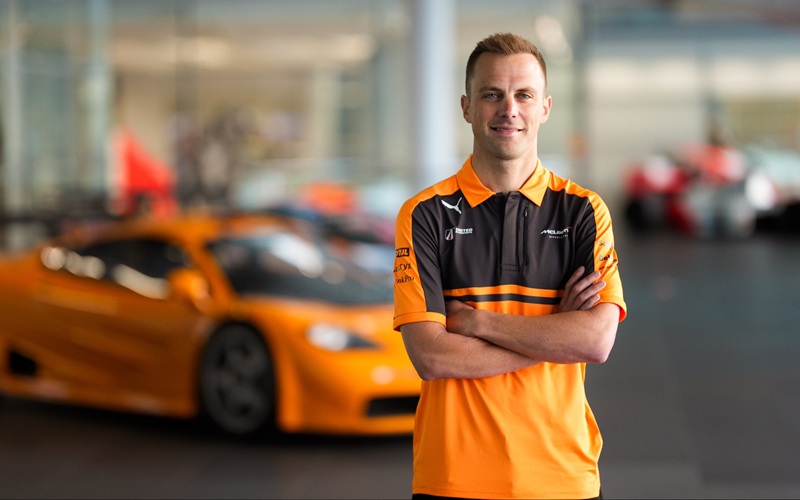 HHyJP2XXgAoyJsy A man in a McLaren polo shirt stands with arms crossed in front of an orange sports car in a modern indoor setting.