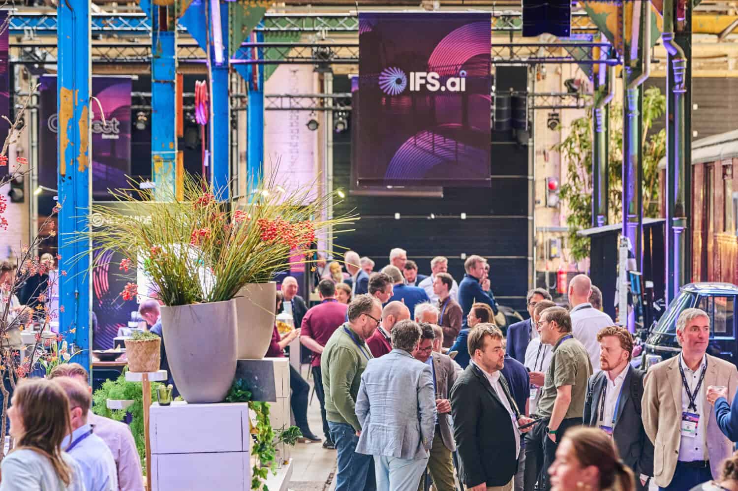 A large group of people in business attire attend a networking event in an industrial-style venue, with IFS.ai and IFS Nexus Black banners hanging above.