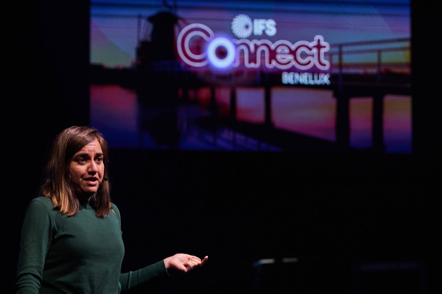 A woman speaks on stage in front of a screen displaying the IFS Connect Benelux logo with a sunset background, featuring the bold design of IFS Nexus Black.