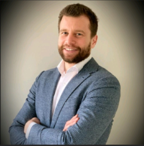Man with brown hair and beard wearing a light blue checked blazer and white shirt stands with arms crossed, smiling at the camera against a plain light background.